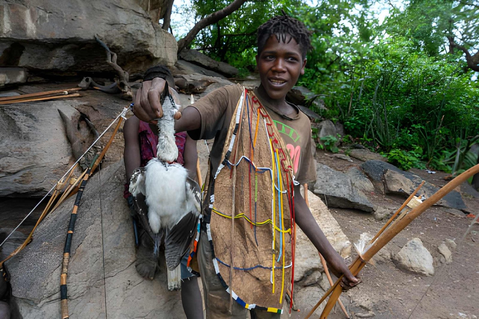 Hadzabe hunter dressed in traditional attire holding a bow and arrow in a wooded forest setting.