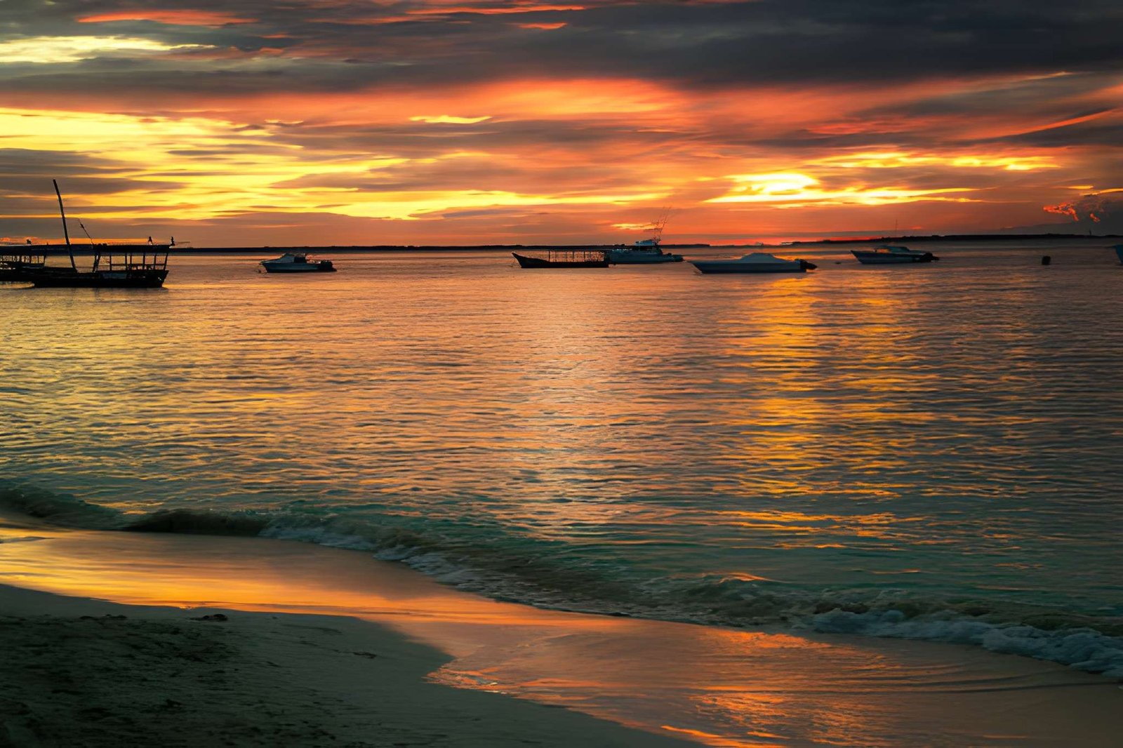 Traditional wooden dhow boats floating on calm waters near a sandy beach during a vibrant golden sunset.