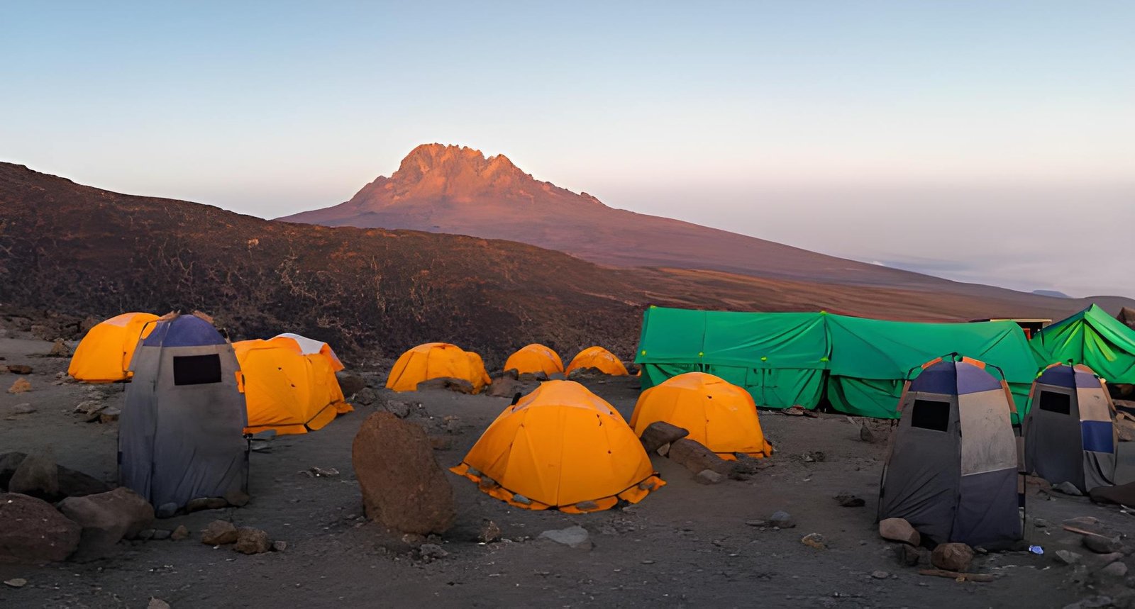 A campsite with bright orange tents set up on a high alpine ridge near a rocky mountain peak.